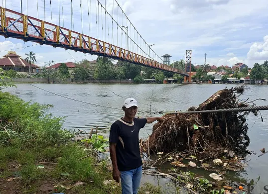 Salah seorang warga, Napong menunjukan pohon rengas yang tumpang dan menimpa kabel baja sling jembatan Setu Tujuh Muara Bojongsari, Kota Depok, Sabtu, 13 Desember 2025. (Sumber: POSKOTA | Foto: Angga Pahlevi)