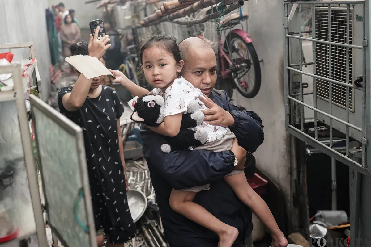 Warga membawa anaknya melewati kebakaran yang menghanguskan pemukiman padat penduduk di kawasan Grogol Utara, Kebayoran Lama, Jakarta Selatan, Minggu, 20 Juli 2025. (Sumber: Poskota/Bilal Nugraha Ginanjar)