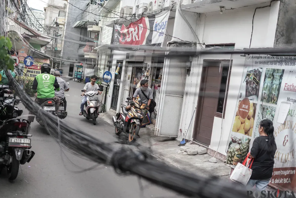 Warga melintas di dekat jaringan utilitas yang menjuntai di Jalan Anggrek II, Karet Kuningan, Setiabudi, Jakarta, Selasa, 11 November 2025. (Sumber: Poskota/Bilal Nugraha Ginanjar)