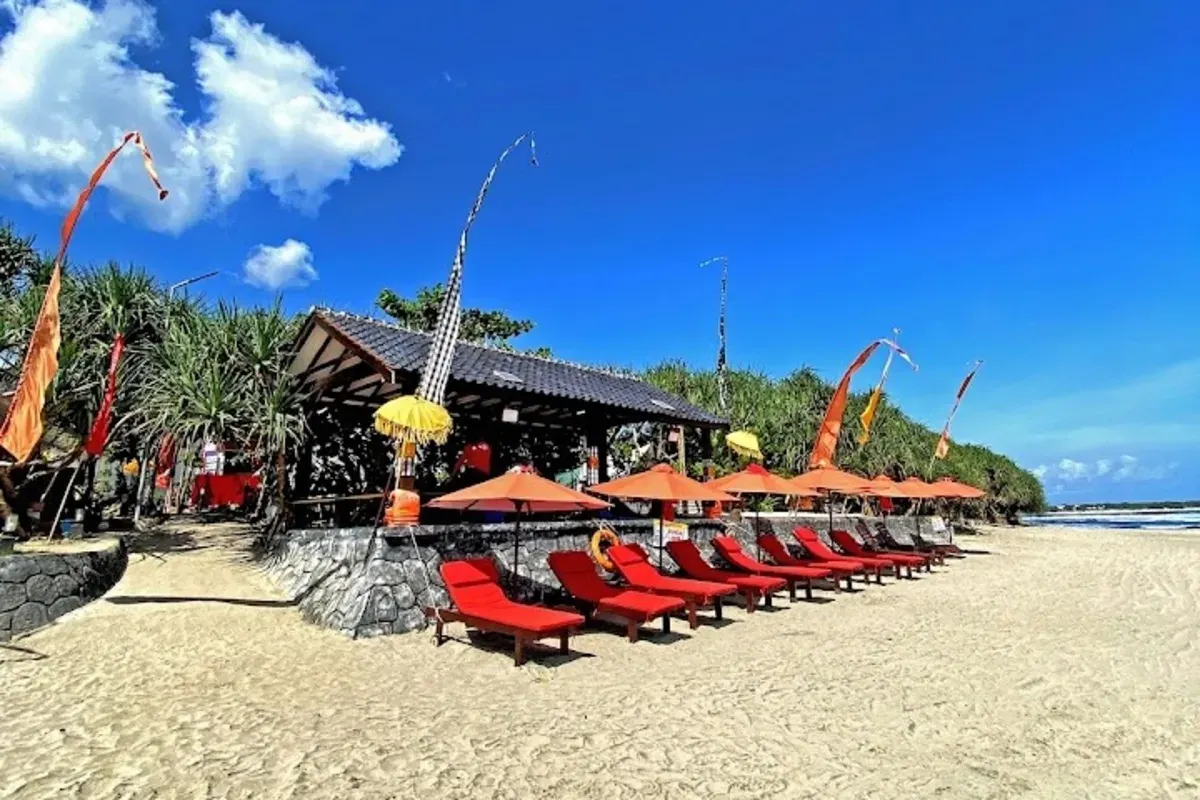 Deretan kursi panjang berwarna merah dengan payung oranye berjajar rapi di atas pasir Pantai Karang Papak, Garut. Nuansa Bali semakin terasa dengan hiasan kain poleng dan umbul-umbul di sekitar area. (Sumber: Villa&Cafe Villa Rahayu Karangpapak)