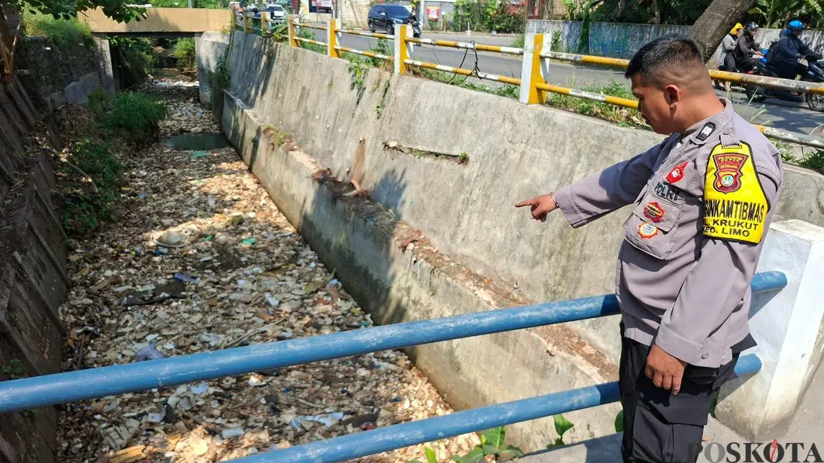 Anggota Bhabinkamtibmas Kelurahan Krukut Aiptu Andriyanto menunjukan lokasi sampah menutup aliran Kali Irigasi RW 03, Jalan Raya Krukut, Limo, Kota Depok, Senin, 13 Juli 2026. (Sumber: Poskota/Angga Pahlevi)