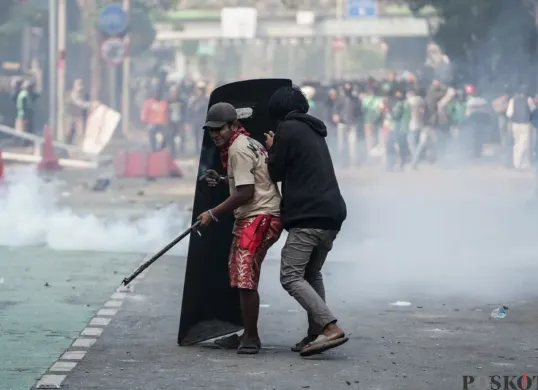 Dua massa aksi berlindung di balik tameng polisi saat demonstrasi di Mako Brimob Polda Metro Jaya, Kecamatan Senen, Jakarta Pusat, Jumat, 29 Agustus 2025. (Sumber: Poskota/Bilal Nugraha Ginanjar)