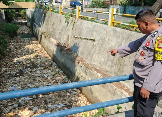 Anggota Bhabinkamtibmas Kelurahan Krukut Aiptu Andriyanto menunjukan lokasi sampah menutup aliran Kali Irigasi RW 03, Jalan Raya Krukut, Limo, Kota Depok, Senin, 13 Juli 2026. (Sumber: Poskota/Angga Pahlevi)